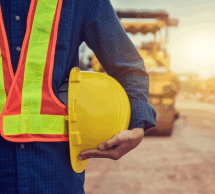 A close-up of a man holding a hard hat at his side, in a construction zone