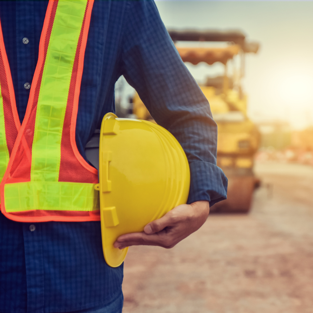 A close-up of a man holding a hard hat at his side, in a construction zone