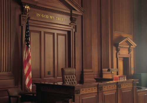 Inside a courtroom looking at the judge's seat, with an American flag in the background and In God We Trust enshrined on the wall